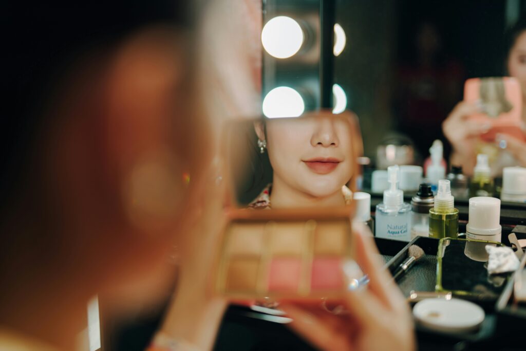 A woman applying makeup reflected in a mirror under bright vanity lights indoors.